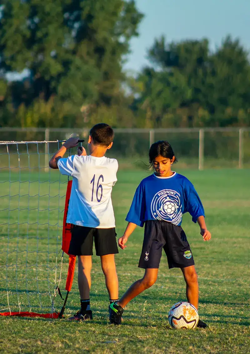 A young girl kicking a soccer ball while a young boy fixes a practice net